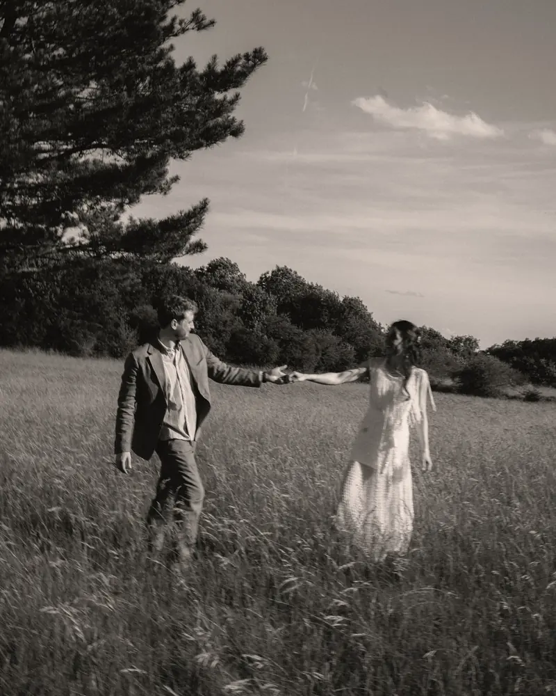 Un homme et une femme en robe blanche marchent main dans la main dans les hautes herbes d'un pré, en noir et blanc