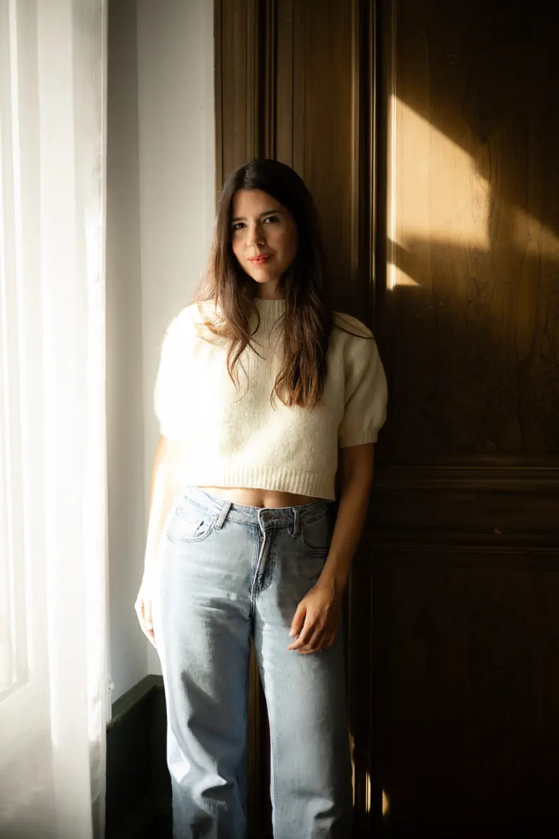 Léa de Nonancourt, pull crème et jean, adossée à une armoire en bois sombre, un rai de soleil sur le mur
