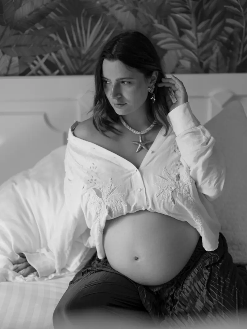Femme enceinte en noir et blanc, assise sur un lit, chemise blanche ouverte sur son ventre rond