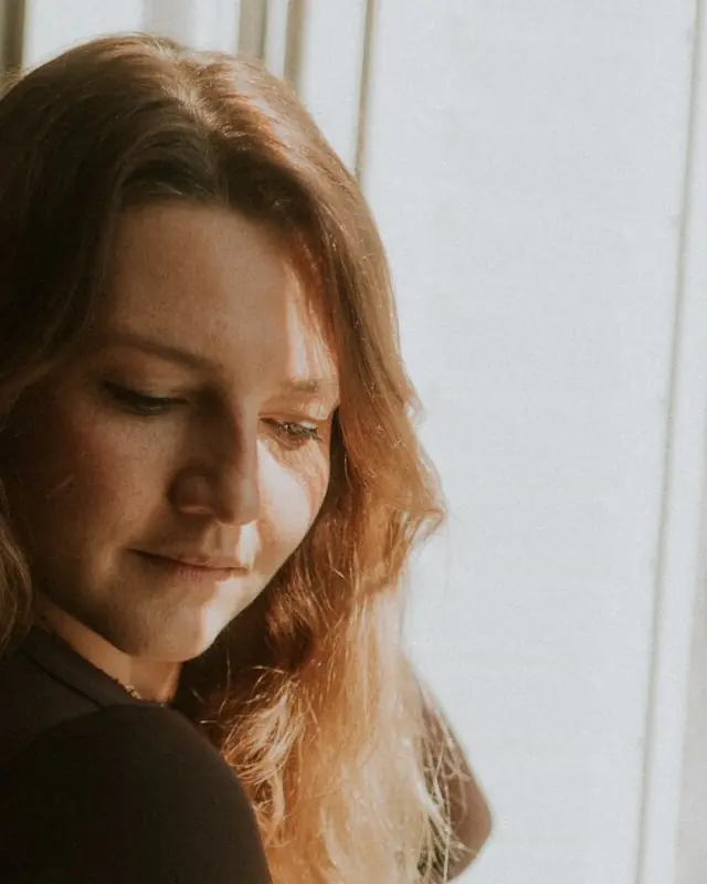Une femme sourit doucement, les yeux baissés, la lumière de la fenêtre sur ses cheveux